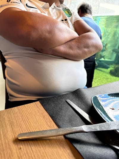 The image depicts an indoor scene at a dining establishment in Mora, Portugal. A seated individual, observed from the chest to mid-thigh, occupies the midground. This person wears a white short-sleeved polo shirt featuring a "SPORTING PORTUGAL" emblem on the left chest and "CITY PAINT" on the right shoulder. Their arms are crossed over a visibly distended abdomen, causing the shirt to stretch tightly. The skin on the visible forearms appears tanned. In the foreground, a wooden table surface transitions to a dark gray placemat. A metallic knife rests diagonally on the placemat, partially obscuring a metallic fork. A square, dark blue and green ceramic plate with minor food remnants is visible on the right side of the placemat. In the background, a second person with gray hair is partially visible from behind, wearing a blue shirt or jacket. To the right of this individual, a large aquarium is present, displaying water with a green tint and multiple dark-colored fish.