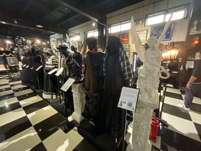 The image depicts an interior scene, possibly a museum or exhibition, with a striking black and white checkered floor. In the foreground and midground, mannequins are displayed, dressed in elaborate, dark-colored historical or theatrical costumes, some with ornate detailing and headwear. One mannequin on the right is dressed in a dark plaid garment and has a black hat with a feather. To the left of this mannequin, a white statue of a winged figure, possibly an angel, stands on a decorative pedestal.

Further into the scene, more mannequins are visible, some wearing dark, flowing dresses and hoods, and one with a fur collar and a colorful scarf. The background shows framed artwork on the walls and display cases with various objects. Lighting fixtures are visible on the ceiling.

A person is partially visible on the far right of the image, wearing jeans and white sneakers, walking away from the camera. The overall atmosphere suggests a curated display of historical attire or artistic presentation.

The prompt mentions the city of Voskhod, Russia. However, there are no visual elements in the image that definitively place it in Voskhod, Russia, or any specific city. The description is based solely on the visual content of the photograph.