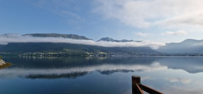 The image depicts a panoramic view of a tranquil fjord in Ørsta, Norway, on a clear day with scattered clouds. In the foreground, a wooden railing and a post are partially visible on the right side of the frame, jutting out into the calm, reflective water. The water mirrors the sky and the distant mountains with remarkable clarity, showing ripples that disrupt the perfect reflection.

The middle ground is dominated by the sprawling town of Ørsta, nestled at the base of imposing, verdant mountains. The town is characterized by a collection of buildings with light-colored roofs, interspersed with patches of green trees. The slopes of the mountains are covered in dense, dark green forests, rising steeply towards the sky. A significant feature is the band of white clouds that elegantly drapes across the mid-section of the mountains, partially obscuring their peaks and adding a dramatic visual element.

The background consists of a clear blue sky, with wispy white clouds scattered across it. The overall impression is one of serenity and natural beauty, showcasing the picturesque landscape of the Norwegian coast. There are no people or animals visible in the image.