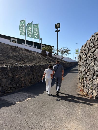 The image depicts a pathway leading towards a McDonald's restaurant and a BP gas station in Antigua, Spain. Two individuals, a man and a woman, are walking away from the camera, hand in hand, down the paved path. The woman is wearing a white outfit, and the man is in a grey outfit. They are casting long shadows on the path due to the bright sunlight.

On the left side of the path, a stone wall separates the pathway from a sloped area with a McDonald's restaurant. The McDonald's building is visible with its signage and three green flags displaying the McDonald's logo. A tall pole with a McDonald's sign is also visible in the distance.

On the right side of the path, a tall stone wall runs alongside the pathway. Further down the path, a BP gas station sign is visible, along with traffic lights. The environment appears to be arid and sunny, with sparse vegetation and palm trees visible in the background. The pathway is asphalted, and there is a manhole cover embedded in the path.