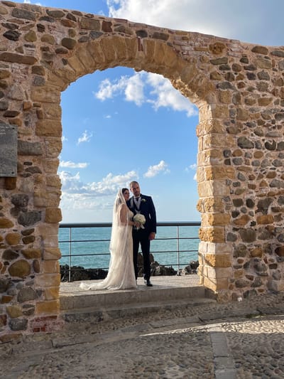 An outdoor, eye-level photograph depicts a bridal couple posing within the Porta Pescara (also known as Porta di Mare or Porta della Marina) archway in Cefalù, Sicily, Italy.
The man, wearing a dark suit, vest, and tie, stands on the right, his left arm linked with the woman's right arm. He has short dark hair and a beard, looking towards the viewer with a neutral expression. The woman, positioned on the left, wears a long, white, beaded wedding gown with a flowing, sheer veil. She holds a white floral bouquet in her left hand and looks towards the man with a faint smile.
They stand centered within the stone archway, which is constructed from irregular, varied-colored, rough-hewn stones, with a distinct keystone. A dark, rectangular plaque with text is partially visible on the inner left wall of the archway. Beyond the couple and through the archway, the bright blue Tyrrhenian Sea extends to the horizon, under a blue sky with scattered white clouds. A dark metal railing with horizontal bars separates the archway's platform from the sea, with dark, jagged rocks visible at the waterline below. In the foreground, a cobblestone street is visible, leading up to the archway via two stone steps. The scene is illuminated by bright daylight.