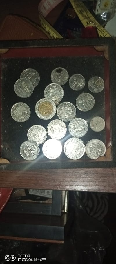 The image displays a collection of coins laid out on a dark surface, possibly a velvet-lined tray or box. The coins appear to be old and vary in size and design. Some coins show visible inscriptions and imagery, suggesting they might be commemorative or historical currency. In the background, a measuring tape with markings is visible, along with a brown leather object, possibly a wallet or pouch. The overall lighting is dim, creating shadows and giving the scene a somewhat muted appearance. There are no people or discernible actions in the image, and the location within the city of Tahmach, Uzbekistan, is not indicated. The scene appears to be a still life arrangement of coins.