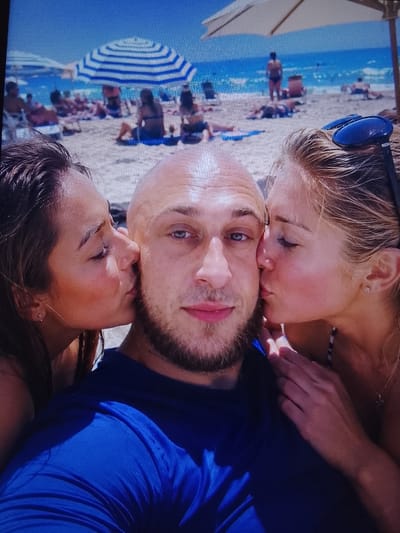 A man with a shaved head and a beard is being kissed on both cheeks by two women. He is wearing a blue t-shirt. The woman on the left has dark hair and is wearing a small earring. The woman on the right has blonde hair and is wearing sunglasses on her head, a necklace, and a striped bikini top. The background shows a beach with people, beach umbrellas, and the ocean. The image is a close-up selfie with a shallow depth of field, making the background blurry. The location is not specified as Safonovo, Russia.