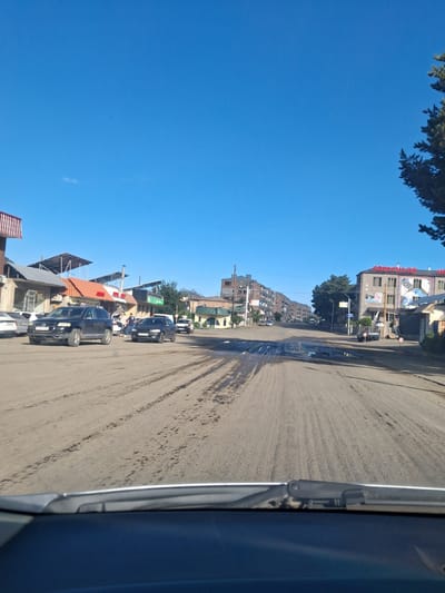 This is a medium-shot, eye-level, outdoor, daytime photograph taken from within a vehicle, looking forward at a street scene in Vayk, Armenia. The road surface is a mixture of dry, dusty dirt and patches of dark, wet mud, with visible tire tracks. On the left side of the road, a black SUV with license plate "37 UK 397" is parked. Further down the street, other vehicles are visible, including a black sedan with license plate "83 4P 316" moving away from the camera, and a small white car. Buildings line both sides of the street. On the left, there are several commercial establishments with awnings and signs, including one with a red roof. On the right, a multi-story building with a red sign that appears to be in Armenian script is prominent, along with other structures. Trees are present on the right side of the image, and the sky is a clear, bright blue. The overall scene depicts a somewhat dusty and unpaved street in a town.