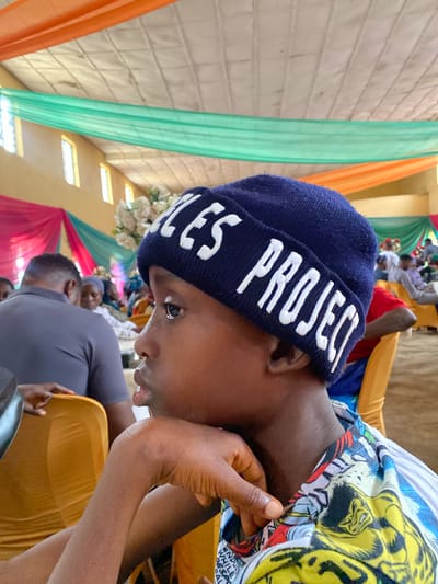 A medium shot captures a young person wearing a blue beanie with white lettering that reads "LES PROJECT," resting their chin on their hand and looking off to the side. The beanie's fabric has a textured knit pattern. The person's face is in profile, with a portion of their ear and neck visible. They are wearing a colorful shirt with a comic book-like print featuring yellow, blue, and red elements. The background suggests an indoor gathering, possibly a church hall or community center in Turu, Nigeria. The ceiling is adorned with orange and green draped fabric, and yellow windows are visible on the wall. Several other individuals are seated in the background, with some partially obscured by chairs. The overall lighting is ambient, with a slight overhead light source. The scene is framed from a slightly low angle, emphasizing the young person as the central subject.