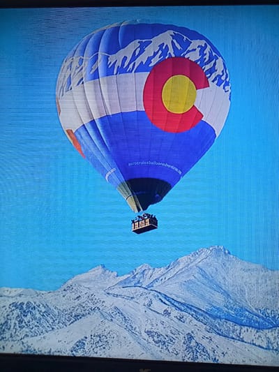 A hot air balloon, decorated with the Colorado flag design featuring white mountain peaks, a red circle, and a yellow circle against a blue background, floats in a clear blue sky. Below the balloon, a snow-covered mountain range is visible. The basket of the hot air balloon carries several passengers. A website address, "astrocruiseballoonadventures.com," is printed on the balloon. The scene appears to be a digitally altered image or a photograph of a screen displaying such an image, given the visible pixelation and screen artifacts. There is no indication of this image being located in Litomyšl, Czechia.