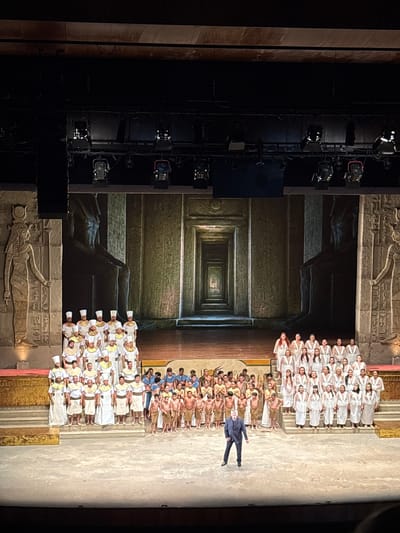 A stage production features a large cast. Some wear white robes and headdresses, while others are bare-chested with similar headwear. A person in a dark suit is centrally positioned, lying horizontally on the stage. The backdrop is a projection or painted scene resembling an ancient Egyptian temple interior with hieroglyphs. There is a tower structure on the left side with theatrical lighting. There is no information indicating that the location is in San Borja, Peru.