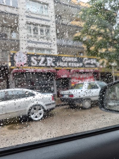 The image captures a street scene in Yenimahalle, Turkey, viewed through a rain-streaked car window. The foreground shows the textured interior of a car door and the edge of the car's window frame. The window itself is covered with numerous raindrops of varying sizes, distorting the view of the outside world.

Beyond the rain-covered window, a multi-story building with light-colored facades and numerous windows is visible. A sign with the text "SZAR" is partially obscured by the raindrops and the reflections. Red awnings or building elements are present below the sign.

Two silver cars are parked on the street. The car closer to the viewer is partially visible, showing its side and a distinctive wheel with multiple spokes. Another silver car is parked behind it. Trees with green foliage can be seen in the background, their colors softened by the rain and distance. The overall atmosphere is one of a rainy day in an urban environment.