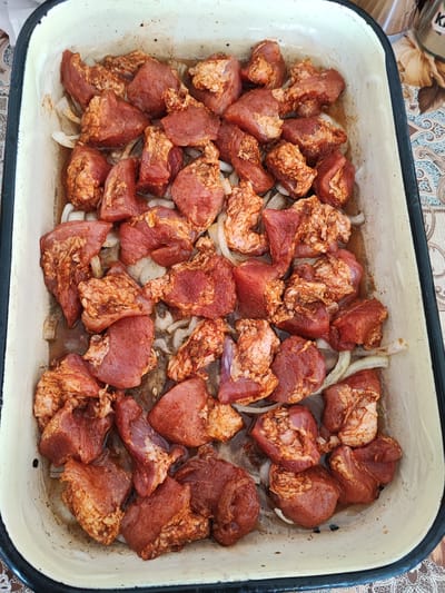 The image shows a rectangular metal baking dish containing raw, marinated meat chunks and sliced onions. The marinade appears to be reddish-brown. The dish is sitting on a surface with a patterned tablecloth or mat. The location is likely Musachevo, Bulgaria. The scene is a kitchen setting with raw food being prepared.