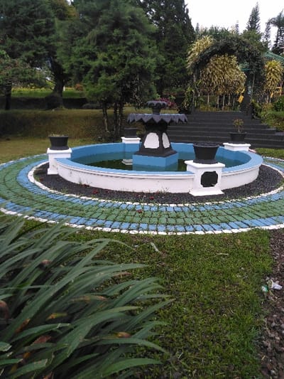 This image presents an outdoor garden scene within the city of Chan-Nua, Laos. A circular fountain basin, with a white exterior wall, occupies the midground and contains light blue-hued water reflecting the overcast sky. A multi-tiered, ornamental structure, predominantly black with white accents on its base, rises from the fountain's center. Four white square pedestals, each topped with a black planter pot, are symmetrically placed around the basin's rim; one pot on the right contains a small plant. Surrounding the fountain is a circular pathway composed of interlocking blue and green square pavers, bordered by a white inner line and an outer ring of black pebbles.
The foreground on the left features a cluster of long, dark green, strap-like foliage. The foreground on the right shows sparse green grass mixed with dark soil and a small piece of white and green litter. In the background, an expansive green lawn and several large, dark green coniferous trees are visible on the left. To the right, a set of dark gray steps ascends beneath an archway covered in dense green foliage, leading to higher garden sections with varied trees, some exhibiting yellowish foliage, and a glimpse of a green roof structure at the far right. The sky overhead is uniformly overcast. No individuals or active subjects are present in the scene.