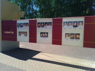 The image shows a bright, outdoor scene with a large, maroon-colored display wall that features several framed photographs of individuals. The wall is positioned in front of lush green trees. The photographs are arranged in sections, with some sections displaying rows of individual portraits and others featuring group photos. The text "ЮНЫЕ ТАЛАНТЫ" (Young Talents) is visible in gold lettering on the left side of the display wall. The ground in front of the wall is paved with light-colored rectangular bricks, and a long shadow is cast across the paving stones, indicating the sun is shining. A portion of a light yellow building or structure is visible on the left side of the image. The overall setting appears to be a public space, possibly a park or an educational institution, given the nature of the display. The image was captured in the city of Otradny, Russia.