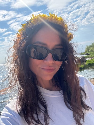 The image shows a woman wearing a dandelion flower crown and sunglasses, with a body of water and vegetation visible in the background. Reflections on the water suggest a sunny day. The sunglasses reflect the surrounding environment, which may include trees and sky. Based on the provided location, Lisy Nos, Russia, this likely portrays a natural setting along the coast or a body of water near this city. The woman is wearing a white t-shirt. The sky behind her contains scattered white clouds.