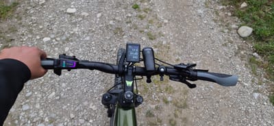 A first-person overhead view depicts the handlebars of an electric mountain bike on a gravel path in Erl, Austria. The rider's left hand, clad in a black long-sleeved garment, grips the left handle. Mounted on the left handlebar is a Bosch Purion controller displaying four active blue battery indicator lights and a purple light indicating the current assistance mode. Centrally mounted on the stem is a Bosch Kiox display showing "65%" battery charge, "eMTB" mode selected, "STRECKE" (trip distance) of 17.0 km, "FAHRZEIT" (ride time) of 01:49, and "GESAMTSTRECKE" (total distance) of 242 km, with a Bosch logo at the bottom. To the right of the display, a black Fischer-branded bicycle headlight is visible, along with a silver and black bicycle bell. The right handlebar features a grey ergonomic grip, a brake lever, and a gear shifter indicating numbers up to '11'. The bike's stem is black with a green 'A' logo and "axess bicycles Designed in Germany" text. The frame visible beneath the handlebars is dark green. The path beneath the bike consists of numerous light-colored gravel stones and dirt, with patches of green grass and a larger, smooth light-colored stone visible on the right edge of the frame.