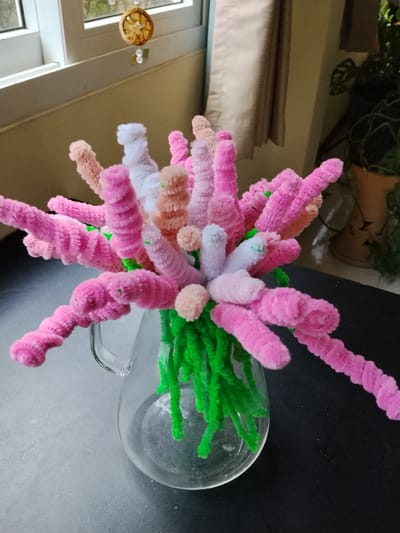 A close-up, eye-level shot shows a bouquet of pipe cleaner flowers in a clear glass pitcher, placed on a dark surface. The flowers are various shades of pink and peach, with some white accents, and are arranged with green pipe cleaner stems. The pitcher is partially filled with what appears to be water. In the background, a window with white trim is visible, with a dried orange slice hanging from what looks like a mobile above it. To the right of the window, a tan curtain hangs, and further to the right, a potted plant with large green leaves is visible in a terracotta pot. The overall lighting suggests an indoor setting, possibly a home or studio. The image's composition and focus are centered on the pipe cleaner flowers, creating a whimsical and colorful display. There is no discernible connection to Ban Patan, Thailand.