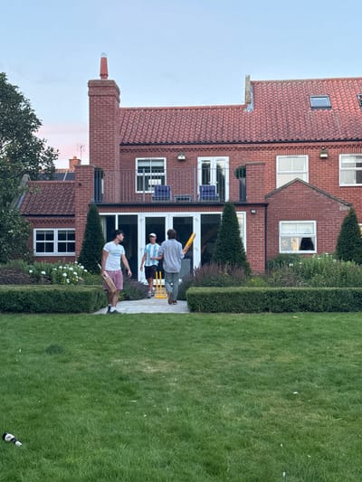 This image depicts a residential garden scene in Bubwith, United Kingdom, captured during the late afternoon or early evening under a clear sky with a faint pink hue near the horizon.
In the background, a large, two-story red-brick house with a red tiled roof dominates the frame. The house features multiple white-framed windows, a prominent brick chimney on the left with a reddish-orange cap, and a second-floor balcony accessible via white double doors. Two blue chairs are visible on the balcony. A skylight is present on the roof to the right. The ground floor incorporates large glass panels, likely patio doors or windows.
The midground features a meticulously maintained garden with a central paved pathway leading towards the house from a large, verdant lawn in the foreground. The pathway is flanked by dark green, neatly trimmed hedges. Behind the hedges, cultivated garden beds contain various green plants and scattered white flowers. Two tall, conical evergreen shrubs are positioned symmetrically closer to the house, flanking the entrance to the paved path.
Three adult males are present in the midground, on or near the paved pathway, appearing to be engaged in or concluding a game of cricket.
- The individual on the left, facing right, wears a white t-shirt with a graphic design, light-colored shorts, and dark shoes. He holds a wooden cricket bat in his right hand, pointed downwards.
- The central individual, facing generally forward, wears a light blue and white striped shirt, a white cap, and dark shorts. He stands next to a set of yellow plastic cricket stumps, complete with bails, placed on the paved surface.
- The individual on the right, with his back mostly to the camera, wears a light-colored long-sleeved shirt and light-colored trousers. He holds a yellow plastic cricket bat horizontally across his body with both hands and appears to be walking away from the house.
In the bottom-left foreground, a white bottle with a dark cap lies horizontally on the green lawn. The lawn itself shows some scattered light-colored debris.