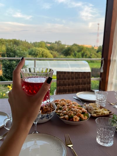A person's hand with black nail polish is holding a glass of red liquid, possibly wine or juice, in the foreground. The glass is made of clear, textured glass. The hand is positioned to the left of the frame. A table is set for a meal with several dishes, plates, glasses, and cutlery. One dish contains a shrimp salad with cucumbers and tomatoes, another contains what appears to be fried mushrooms, and a third contains pieces of cheese and possibly some other food items. The table is covered with a mauve tablecloth. In the background, a balcony railing is visible, beyond which is a lush green landscape with trees and a greenhouse structure. The sky is partly cloudy with a pale blue hue. The scene appears to be an outdoor dining setting, likely on a porch or balcony, with a view of a rural or semi-rural environment. The overall impression is one of a relaxed meal being enjoyed outdoors, possibly in the late afternoon or early evening. The location is identified as Суроватиха, Russia.