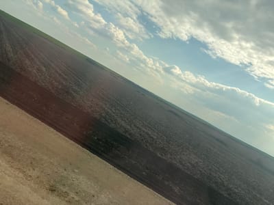 The image depicts an agricultural landscape observed from an oblique angle, situated in the city of Svoboda, Bulgaria. In the lower left foreground, a strip of light brown, uncultivated earth or a dirt path runs diagonally. This strip transitions into a vast expanse of dark brown, tilled agricultural land, exhibiting discernible parallel furrows or crop rows that extend across the midground. Beyond the tilled field, a narrow band of vibrant green vegetation or crops is visible along the distant horizon. The upper right background features a partly cloudy sky, displaying scattered white and grey cumulus formations against patches of clear blue. No individuals, structures, or identifiable machinery are present within the scene.