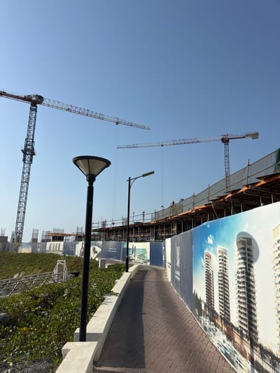 This media file depicts an outdoor scene under a clear blue sky, suggesting a bright day, likely morning or afternoon, given the clear visibility and lack of harsh shadows. The image is oriented vertically, with the sky dominating the upper portion and the ground plane at the bottom.
The primary subjects are several construction cranes, characteristic of a building site. The cranes are large, metal structures with intricate lattice frameworks, some extending high into the sky. One crane prominently displays the "RAIMONDI" brand name. In the foreground, a modern lamppost with a distinctive wide, conical shade is visible. Behind the lamppost, a paved walkway or path runs diagonally across the frame, bordered by a low concrete wall. Abundant green vegetation, possibly ground cover or low-lying shrubs, grows alongside the path and in other areas of the scene.
The background reveals the ongoing construction of a building, with scaffolding and partially completed structures. A large, illustrative banner is affixed to the construction site, showcasing a rendering of sleek, modern high-rise buildings against a sky with clouds, hinting at the future development of the area. The banner also contains text, although it is partially obscured and difficult to read clearly in its entirety. Some visible text includes "AL JAHRA WATERFRONT."
The overall impression is one of active development and urban progress. The scene is devoid of people, focusing instead on the industrial and architectural elements of the construction project. The setting appears to be a public area near a waterfront, given the mention of "WATERFRONT" on the banner. The image conveys a sense of a large-scale project underway in a developing urban or semi-urban environment.