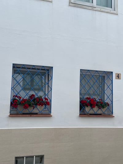 A portion of a building facade in Calella De Palafrugell, Spain, is depicted. The upper section of the facade consists of white textured stucco, while the lower section features light beige rectangular tiles. Two identical, square windows are visible in the middle section, each fitted with a blue metal security grille forming a diagonal grid pattern. Below each window, a terracotta-colored sill supports a rectangular, light-colored planter box. Both planter boxes contain vibrant red geraniums and green foliage. A small, square ceramic tile bearing the black numeral '4' is affixed to the white stucco wall to the right of the rightmost window. Partial views of additional windows are present at the top and bottom edges of the frame. No individuals or discernible actions are depicted in the scene.
