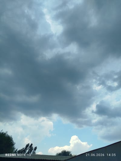 A wide-angle, medium-altitude shot captures a dramatic sky filled with a mixture of dark, heavy clouds and patches of bright blue sky. The clouds are predominantly gray and overcast, suggesting a stormy or pre-storm condition, with some lighter, puffy cumulus clouds visible towards the bottom of the frame. In the foreground, the top edge of a corrugated metal roof is visible, angled slightly upwards from left to right. To the left of the roofline, dark green foliage from trees protrudes into the frame, with some of the leaves appearing blurred, possibly due to wind. The bottom left corner of the image displays white text reading "REDMI NOTE 13". In the bottom right corner, white text indicates the date and time: "21.06.2026 14:35". The scene is set in Асан-Чек, Kyrgyzstan.