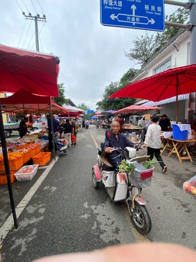 This image captures a street scene in Chongzhou City, China, likely a market given the presence of stalls and produce. An elderly man is seen riding an electric scooter down a paved road lined with red and white awnings, typical of outdoor vendors. The man, wearing a blue jacket and holding the handlebars, looks towards the camera. The scooter is equipped with a basket carrying green vegetables and some red items.

In the background, several market stalls display fruits and vegetables in orange and white crates. Other people are visible walking and browsing, contributing to the bustling atmosphere. A blue street sign in the upper left corner provides location context, with Chinese characters and English translations: "梓潼大道 Zitong Avenue," "裕民街 Yumin Street," and "兴盛街 Xing Sheng Street." The weather appears overcast, with a cloudy sky. The overall scene suggests a lively local market day, with everyday commerce taking place.