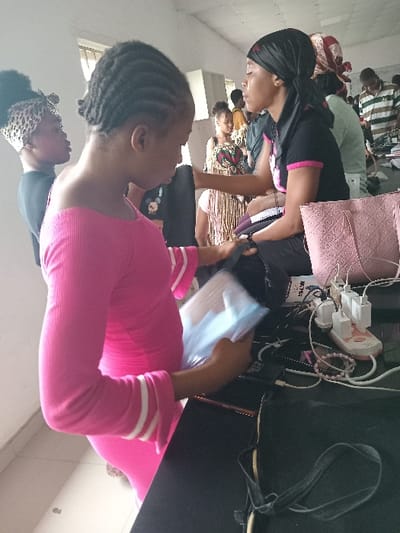 This image depicts an indoor, well-lit communal space in Ogboko, Nigeria. The room has light-colored walls and a window with vertical bars visible in the upper left background.
In the foreground, a female with dark skin and braided hair wears a tight-fitting, off-shoulder pink long-sleeved dress with white stripes on the cuffs. She holds a transparent, light-blue plastic package containing several rectangular items in her right hand, positioned over a dark-surfaced table. To her right, a second female, also with dark skin, wears a black headwrap and a black short-sleeved t-shirt with pink accents. She is seated behind the table, facing left. A purple object rests near her lap.
The dark table surface features a pink textured handbag, a white power strip with multiple white USB charger adapters plugged in, several tangled charging cables (white and black), and a dark elastic headband.
In the mid-ground, to the far left, a female wearing a leopard-print headwrap and black clothing is partially visible. Further back, a female stands in a light-colored dress with horizontal stripes, fringe-like embellishments on the skirt, and a patterned bust. Additional indistinct figures are visible in the background, including one wearing a horizontally striped shirt, indicating a larger gathering of individuals in the space. The central figures appear to be engaged with items on the table, suggesting an organized activity or a workshop setting.