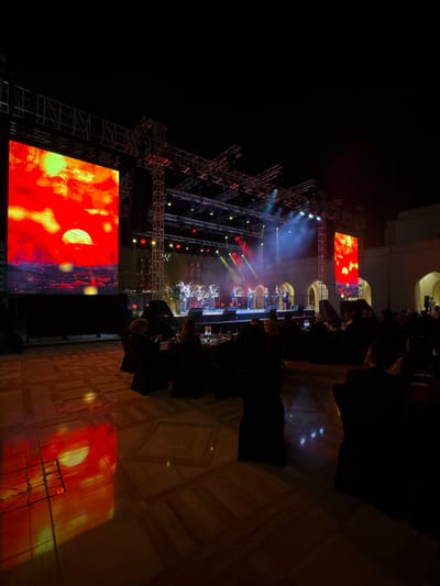 A nighttime concert scene features a stage with lighting rigs and large screens displaying abstract red images. A band performs in the center, illuminated by stage lights and visible through a slight haze. Dark silhouettes of audience members fill the foreground, with a polished floor reflecting some of the stage lights. A building structure is faintly visible in the upper right corner.