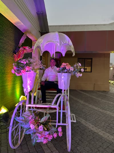 A man sits in a white bicycle carriage decorated with flowers, beneath a white, ornamental umbrella. The scene is bathed in purple and yellow light, with an artificial green lawn beneath the carriage. The background includes building facades with a brick path to the right. The location is likely in Nashik, India, as indicated in the prompt.