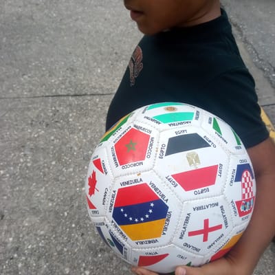 A close-up, over-the-shoulder shot captures a child in a black t-shirt holding a white soccer ball adorned with various national flags and country names. The ball is positioned in the lower right quadrant of the frame, with the child's hands visible supporting it. The background consists of a grey, textured asphalt surface, suggesting an outdoor urban or park setting. The lighting is natural, casting subtle shadows and highlighting the details of the soccer ball, which appears to be a souvenir item featuring representations of flags from countries like Argentina, Morocco, Egypt, Nigeria, Croatia, Venezuela, Canada, England, and Italy. The image composition emphasizes the soccer ball and the child's interaction with it.