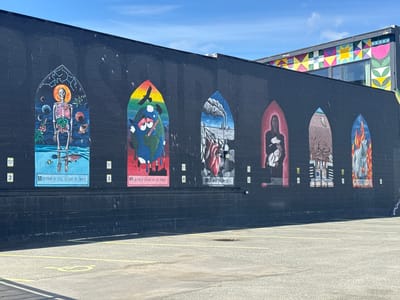 The image captures a mural in Anchorage, Alaska, in the United States of America. The mural is painted on a long, black, brick wall with seven distinct painted sections in the form of arched panels, each showcasing a different piece of art with associated text. The first arch from left to right contains a human skeleton with various planets and organs. The second arch contains the earth with a bird, blood red skies and a rainbow, with the words "OUR ONLY HOME IS ON FIRE". The remaining five arches contain similar themes regarding the environment, with pollution, people, land and animals. Each of the arches is marked with a number starting with 2, increasing up to 8. In front of the mural is an empty parking lot with yellow parking lines and a disabled parking symbol. The sky is clear and blue, and part of a building with a brightly colored design on its side is visible behind the mural.