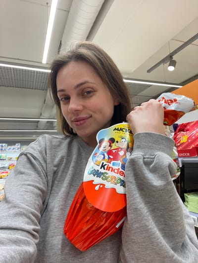 An adult female is depicted in a retail setting, specifically a supermarket aisle in Monsummano Terme, Italy. She possesses light brown hair and a fair complexion, and is wearing a plain grey crew-neck sweatshirt. Her left arm is raised, holding a large Kinder Gran Sorpresa chocolate egg. The egg is enveloped in red foil and features a white and blue cardboard sleeve displaying "Disney Mickey and Friends" characters (Mickey Mouse, Goofy, Donald Duck) along with the brand name. The female is looking directly at the camera with a faint smile. The background consists of white ceiling panels with linear fluorescent lighting fixtures and blurred shelving units stocked with various products, including confectionery like Lindt, indicating a commercial grocery environment.