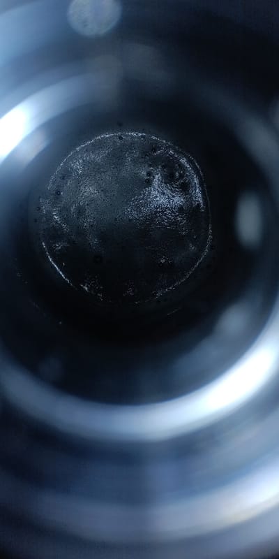 The image shows a close-up, overhead view of the bottom of a dark container, possibly a cup or bottle. The surface is dark and appears to be wet or coated with a dark residue, possibly coffee grounds or sediment, with small bubbles visible. The edges of the container are visible as blurred, concentric rings of light and dark, suggesting a cylindrical interior with a metallic or reflective surface. The lighting is dim, with a bright highlight in the upper left quadrant and a softer, broader highlight on the right side, indicating artificial light sources. There are no people or visible actions. The image does not contain any information about the city of Gornyak, Russia.