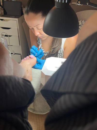 In Montrond-les-Bains, France, a close-up shot captures a woman's hands, adorned with blue gloves, meticulously applying small pink daisy decorations to a person's toenails. The focus is on the detailed nail art process. A white UV lamp, commonly used for curing nail polish, is visible on the table. The scene is illuminated by a black desk lamp, casting light on the subject's work. The background features drawers and other items, suggesting a personal grooming or salon setting. The image is taken from a low angle, with parts of another person's legs in the foreground, creating a sense of intimacy and personal perspective.