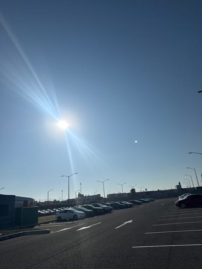The image depicts a bright, sunlit parking lot in an urban environment, potentially located in Sanctorum, Mexico. A row of parked cars lines the right side of the frame, alongside painted parking lines. Streetlights are visible along the length of the parking area. The sky occupies a large portion of the image, with a bright sun near the bottom-left casting intense rays across the scene. The horizon shows silhouettes of buildings or structures, possibly part of the urban landscape. No people are visible in the image.