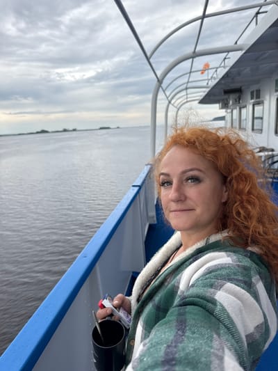 A woman with long, curly red hair is taking a selfie on the deck of a boat. She is wearing a green and white plaid jacket. The boat is on a wide, calm river with land visible in the distance. The sky is overcast. A black mug and a pack of cigarettes are visible in her hand. The location is the city of Zuevo, Russia.