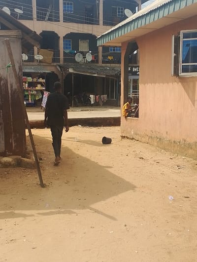 The scene takes place in Ogharefe, Nigeria at a location with the coordinates 5.935 latitude and 5.67 longitude. A man in a black shirt and black pants walks away from the camera on a dirt path in front of a building. The building is made of concrete and has a green roof and a window. There is a black bucket on the ground in front of the building. There is another person sitting on the steps of the building, leaning against the wall, and a third person is sitting on the ground to their right. There is a small, open-air market in the background, behind the walking man. It is daytime and the sun is shining.
