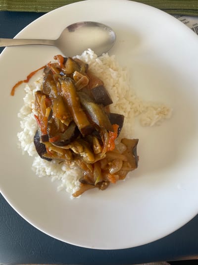 A close-up, overhead shot shows a white, oval plate filled with white rice topped with a serving of cooked eggplant and tomato stew. A silver spoon rests on the upper left side of the plate, partially covering the rice. The plate is set on a dark blue textured surface, possibly a placemat, with a sliver of green fabric visible at the top edge. A small portion of a white paper tag with black text, including the word "Green" and numbers like "15cm" and "0925," is visible on the green fabric. The scene suggests a meal is being served or has just been presented. There is no indication of the city of Bendigo, Australia, or any other specific location.