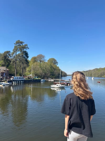 A young person with blonde hair, wearing a black t-shirt and grey leggings, stands by a body of water, likely a river or estuary. The water reflects trees and boats. Several boats are moored along the shoreline. The sky is clear blue. The location is likely within Staffordshire Moorlands, United Kingdom.