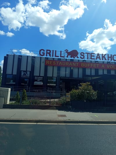 This is a low-angle, outdoor, medium shot of a restaurant with a bright blue sky and white clouds in the background. The restaurant has a large red sign that reads "GRILL STEAKHOUSE RESTAURANT BUFFET A VOL". Below the sign, there are several windows and some greenery. A fence is visible in front of the greenery. A paved road runs alongside the building. The sun appears to be shining, indicated by bright white clouds and the lighting on the building. The image was likely taken during the daytime. Visible text includes "GRILL STEAKHO", "RESTAURANT BUFFET A VOL", "BUFFET A EMPORTER PETITE BARQUETTE 6,00 €", "SALLE CLIMATISÉE", "GRANDE BARQUETTE 11,00 €", and the number "15" on a yellow sign. The setting appears to be a commercial area in Laxou, France. There are no people visible in the image.