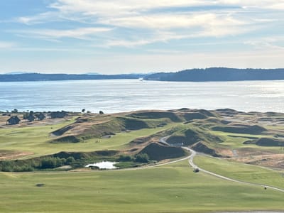 The aerial photograph captures Chambers Bay Golf Course in University Place, Washington, USA. The foreground features meticulously maintained green fairways and tee boxes, with a red flag indicating a hole on the left. A golf cart is parked on a gravel path in the middle ground. Further into the scene, the course transitions into undulating, natural dunes covered in dry grasses and vegetation. Several small, rustic buildings with peaked roofs are visible on a ridge overlooking the course. A pond or water hazard is situated amongst the dunes. The middle ground of the image is dominated by the expansive, shimmering body of water that appears to be Puget Sound, with a distant, tree-covered shoreline forming the horizon under a pale blue sky with scattered white clouds. Several distant boats are visible on the water.
