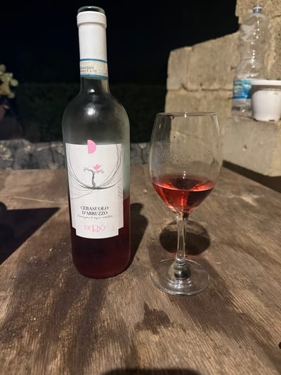 The image displays a bottle of "CERASUOLO D'ABRUZZO" wine and a wine glass filled with rosé wine, both resting on a wooden surface. The bottle has a white label with an artistic drawing of a tree and a pink bird, along with text that reads "CERASUOLO D'ABRUZZO" and "DE RIO". There is also a partially visible plastic bottle and a white dispenser in the background, suggesting an outdoor or rustic setting. The lighting indicates it might be evening or nighttime. The overall mood is relaxed and perhaps social, hinting at a moment of enjoying wine. The provided location context of Carpignano Salentino, Italy, adds a regional flavor to the scene.