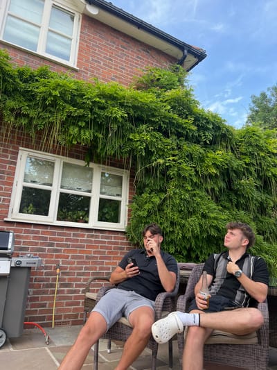 Two young men are sitting on patio furniture. One wears a black and white striped shirt and holds a bottle. The other wears a black polo and grey shorts, using a cell phone. Behind them is a brick house covered in dense green foliage. A white window is visible in the brick wall. A grill is on the patio floor. The sky is visible at the top left.