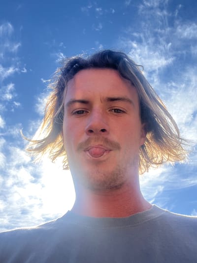 A man with blond hair, wearing a gray t-shirt, is looking down at the camera, sticking his tongue out. The background is a bright blue sky with wispy white clouds. The location is Bellambi, Australia.