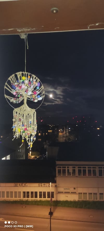 The photograph, taken at night, presents a view from an elevated position overlooking a cityscape. In the lower left foreground, a brown railing is visible. A decorative object, likely homemade, resembling a tree or sun-catcher, hangs suspended by a thin line. It's crafted from twine and adorned with colorful beads.
The background reveals a nighttime urban landscape with varying light sources. Buildings with illuminated windows dot the scene. The sky is dark, with a bright, presumably full, moon partially obscured by clouds. Red lights are scattered across the horizon, possibly indicating towers or other structures.
To the right of the beaded ornament is a building with many windows. Visible graffiti is present on the building.
The image includes a watermark in the bottom right corner, indicating it was taken with a POCO X3 Pro camera.
Location: The building is located in Constance, Germany. The graffiti says "Gött 2.0", which means "God 2.0" and it is believed that it stands for "Gottmannshausen 2.0" in Constance.