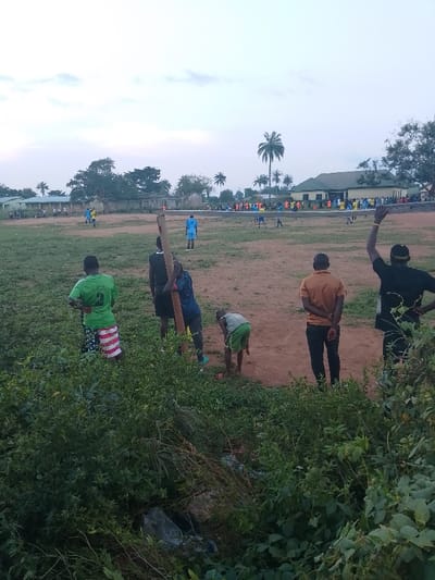 The image depicts an outdoor scene at a football field in Ugwolawo, Nigeria, under an overcast sky. In the foreground, dense green shrubbery and scattered litter occupy the lower portion of the frame. Six individuals are positioned in the foreground, facing towards the field. From left to right, one male wears a green jersey with the number "2" and patterned shorts. Adjacent to him, two males are positioned near a vertically planted wooden pole; one in dark attire leans against it, while another in a blue shirt also stands by it. A young male in a green shirt and shorts is bent over near the pole's base. To their right, a male in an orange shirt and dark trousers stands with hands behind his back. Furthest right, a male in a black shirt raises his right arm.

The midground reveals a large, uneven dirt field with sparse grass. An informal football match is in progress, with multiple players visible across the field. Players in light blue jerseys are seen on the left and center of the field, while a group of players in assorted light blue and yellow/orange jerseys are gathered on the right side of the field near a potential goal area.

The background features several buildings and abundant vegetation. On the far left, a light-colored, single-story building is visible. Towards the center-left, dilapidated concrete structures are present. A larger building with a green roof and light-colored walls stands towards the right of center. Numerous trees, including several tall palm trees, are distributed across the background and along the perimeter of the field.