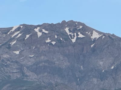 The image shows a mountain range with patches of snow against a clear blue sky. The mountain is rocky and rugged, with sharp peaks. The snow is scattered across the slopes and crevices, suggesting a high altitude or recent snowfall. The lighting indicates it is daytime, likely late afternoon or early morning due to the soft light. The lower part of the image shows some greenery, possibly foothills or a valley floor. The scene is natural and devoid of any man-made structures or human presence. It is not possible to determine the specific location within Uchterek, Uzbekistan based solely on this image.