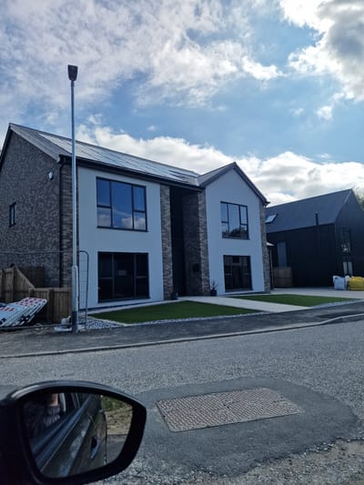 A daytime street view in Witton Gilbert, United Kingdom, shows two modern houses with a clear blue sky and scattered white clouds overhead. The image is taken from what appears to be a car, with a side mirror visible in the bottom right reflecting part of the vehicle's interior.
The house in the foreground features a light-colored rendered upper facade and a darker, possibly brick or stone, lower facade. It has large, rectangular, dark-framed windows and a dark-tiled roof, which appears to be fitted with solar panels. A tall, slender streetlight pole is prominent in the left foreground.
Further down the street, behind the first house, another contemporary house is visible, characterized by its dark cladding and dark roof. The street itself is paved with an asphalt-like material, and a textured metal manhole cover is clearly visible on its surface. Adjacent to the street, there's a narrow strip of gravel and artificial green turf acting as front yards for the houses. Parts of white and red construction barriers are faintly visible towards the lower left. The overall scene suggests a newly developed or recently renovated residential area. No discernible text is visible on any objects.