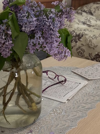 A close-up, eye-level shot captures a bouquet of light purple lilacs in a clear glass jar filled with water. The jar is placed on a light brown wooden surface, possibly a table, covered with a light grey, patterned lace doily. A pair of purple-rimmed glasses rests on top of a folded piece of paper with a grid-like pattern, which is also on the doily. In the background, a blurred, upholstered piece of furniture with a floral pattern is visible. The lighting is soft and natural, suggesting an indoor setting. The image does not contain any identifiable landmarks or features that would indicate its location within the city of Jrarat, Armenia.
