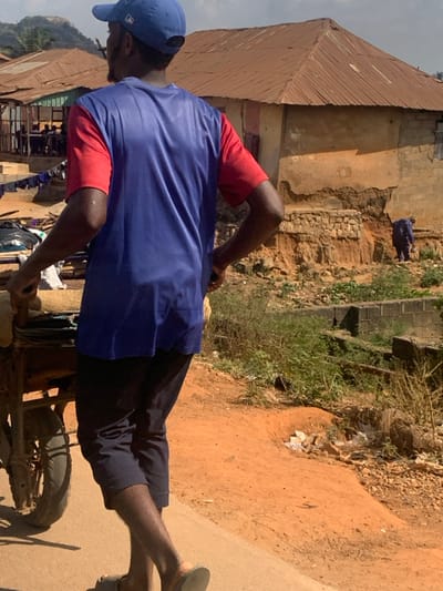The image displays a daylight scene in Egbedi, Nigeria. In the foreground, a dark-skinned male, viewed from the back, walks left while pushing a two-wheeled cart. He wears a blue baseball cap, a blue t-shirt with red short sleeves, dark trousers, and light-colored sandals. The cart, equipped with a dark-colored wheel and tire, carries stacked items, possibly wrapped goods or documents.
The ground is an unpaved reddish-brown dirt path, with a visible concrete or paved edge to the immediate left. To the right of the path, the terrain drops off into an area of disturbed reddish soil and sparse green vegetation.
In the midground, a single-story building with light-brown, textured walls and a rusted, corrugated metal pyramidal roof stands on the right. Its foundation exhibits significant erosion, exposing layers of earth and stonework. A small, dark opening, possibly a window, is visible on its side. Further into this eroded area, a second, less distinct figure, dressed in blue, appears to be stooping or working. To the left, another single-story structure with a similar rusted roof is visible, with laundry hanging on a line nearby.
The background features distant, hazy hills or rock formations beneath a light blue sky with scattered white clouds.