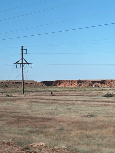 A lone individual rides a dirt bike across a vast, dry, grassy field under a clear blue sky. In the foreground, a large electricity pylon stands tall, with power lines stretching horizontally across the frame. In the background, a rugged, reddish-brown cliff face rises sharply from the terrain. The scene suggests a rural or semi-arid environment, possibly on the outskirts of Ильинка, Russia.